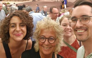 Selfie im Zuschauerraum des Naturtheaters Reutlingen: Beate Müller-Gemmeke steht lächelnd mit drei weiteren Besucher vor der Bühne. Im Hintergrund sind die offene Bühne, das Bühnenbild und bereits einige Zuschauer zu sehen. Die Atmosphäre wirkt freundlich und gelöst.