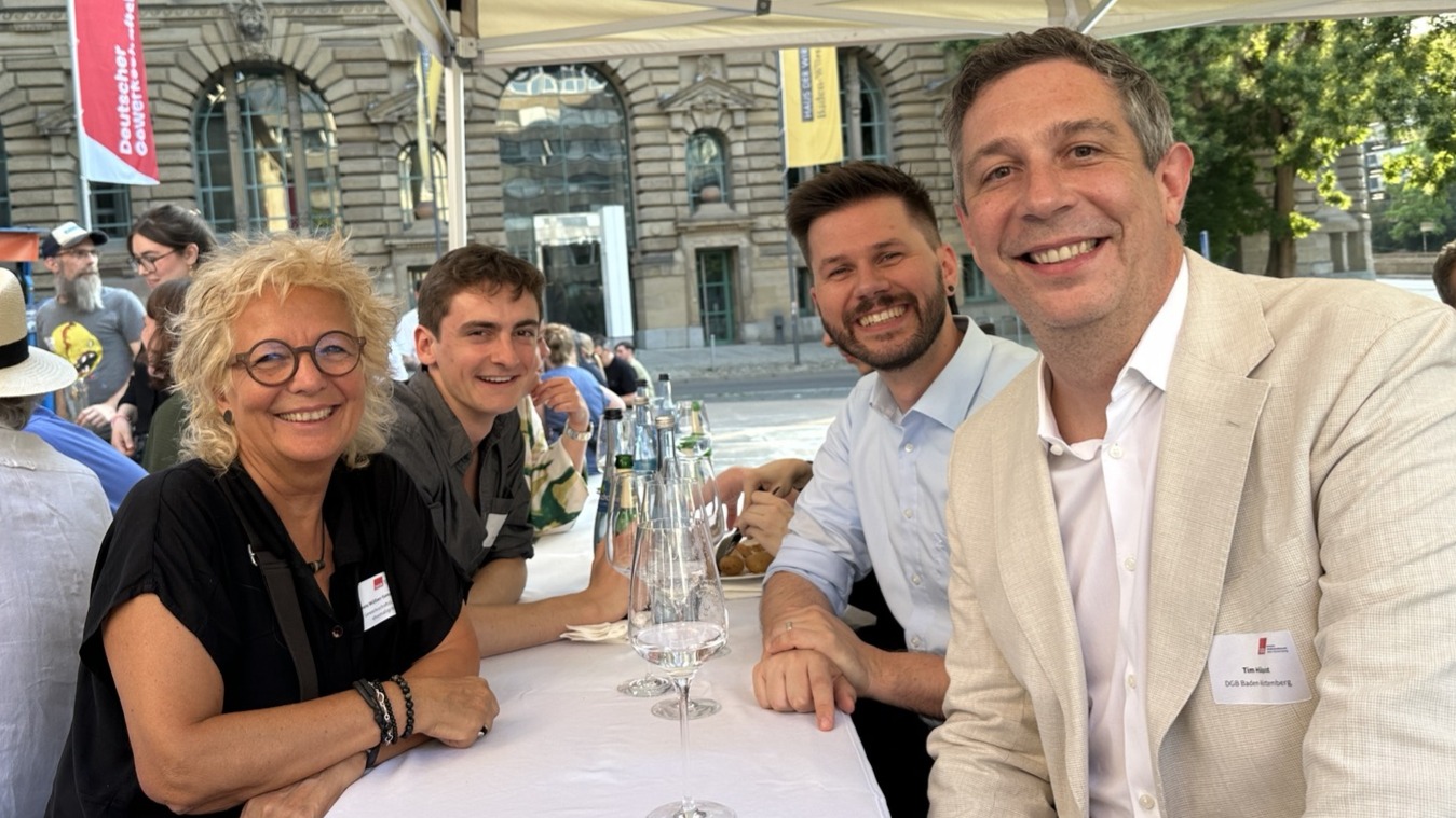 Beate Müller-Gemmeke sitzt beim Sommerempfang des DGB in Stuttgart mit drei weiteren Gästen an einem langen Tisch. Alle lächeln in die Kamera. Im Hintergrund sind weitere Besucherinnen und Besucher, das Stuttgarter Gewerkschaftshaus und eine Fahne des Deutschen Gewerkschaftsbunds zu sehen.
