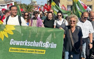 Demonstrationszug beim DGB-Aktionstag in Stuttgart mit vielen Gewerkschaftsfahnen. Im Vordergrund tragen mehrere Teilnehmende ein großes grünes Banner von GewerkschaftsGrün mit der Aufschrift „Gerechte Löhne & soziale Sicherheit“.