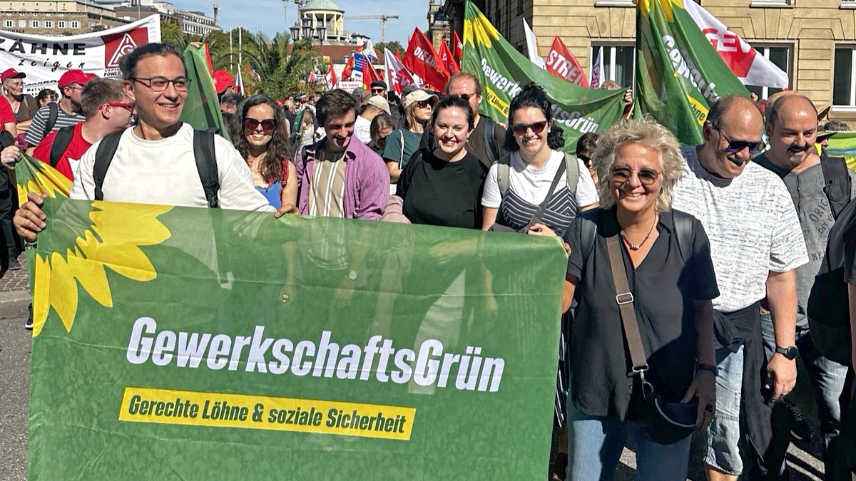 Demonstrationszug beim DGB-Aktionstag in Stuttgart mit vielen Gewerkschaftsfahnen. Im Vordergrund tragen mehrere Teilnehmende ein großes grünes Banner von GewerkschaftsGrün mit der Aufschrift „Gerechte Löhne & soziale Sicherheit“.