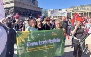 Kundgebung auf dem Stuttgarter Schlossplatz bei sonnigem Wetter. Drei Frauen stehen hinter einem großen Banner von GewerkschaftsGrün, im Hintergrund sind zahlreiche Demonstrierende sowie rote Gewerkschaftsfahnen zu sehen.