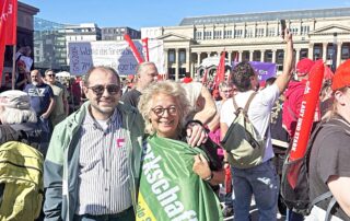 Zwei Teilnehmende stehen gemeinsam auf der Kundgebung in Stuttgart und lächeln in die Kamera. Hinter ihnen sind viele Demonstrierende und Gewerkschaftsfahnen zu sehen, eine grüne GewerkschaftsGrün-Fahne liegt über der Schulter der Frau.