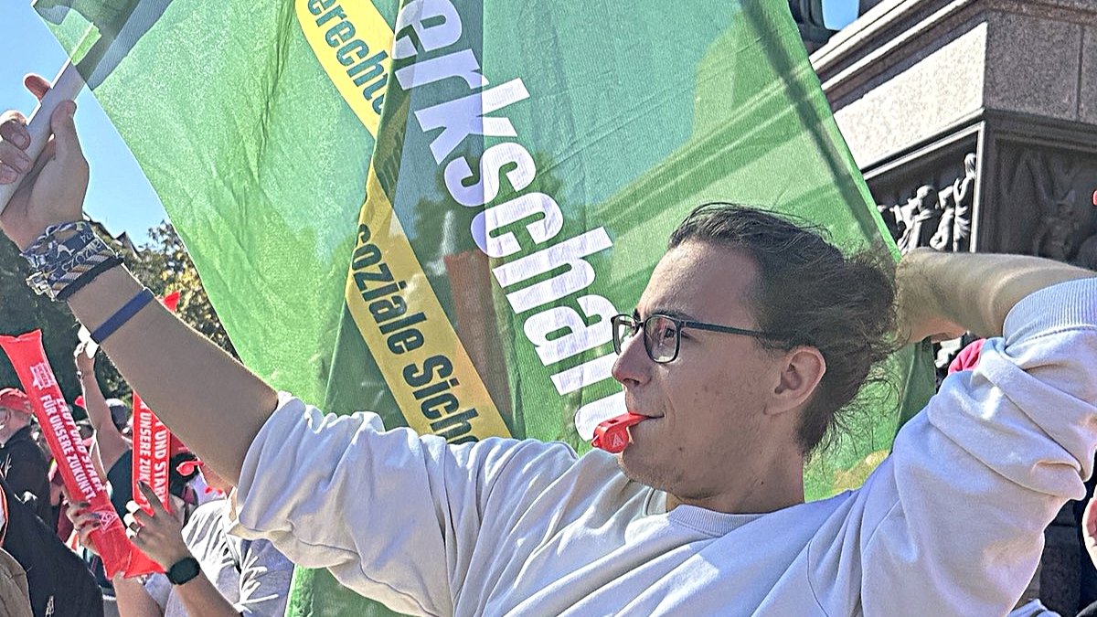 Ein junger Mann macht während der Kundgebung ein Selfie. Hinter ihm weht eine große grüne Fahne von GewerkschaftsGrün mit der Aufschrift „Gerechte Löhne & soziale Sicherheit“.