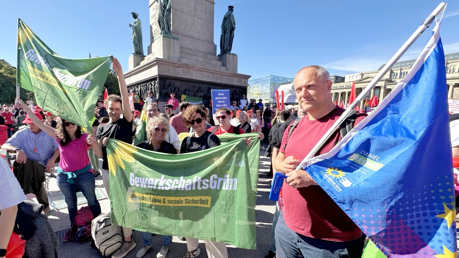 Gruppe von GewerkschaftsGrün beim DGB-Aktionstag auf dem Stuttgarter Schlossplatz. Mehrere Teilnehmende halten ein großes GewerkschaftsGrün-Banner und grüne Fahnen, ringsum stehen viele weitere Demonstrierende mit Gewerkschaftsfahnen.
