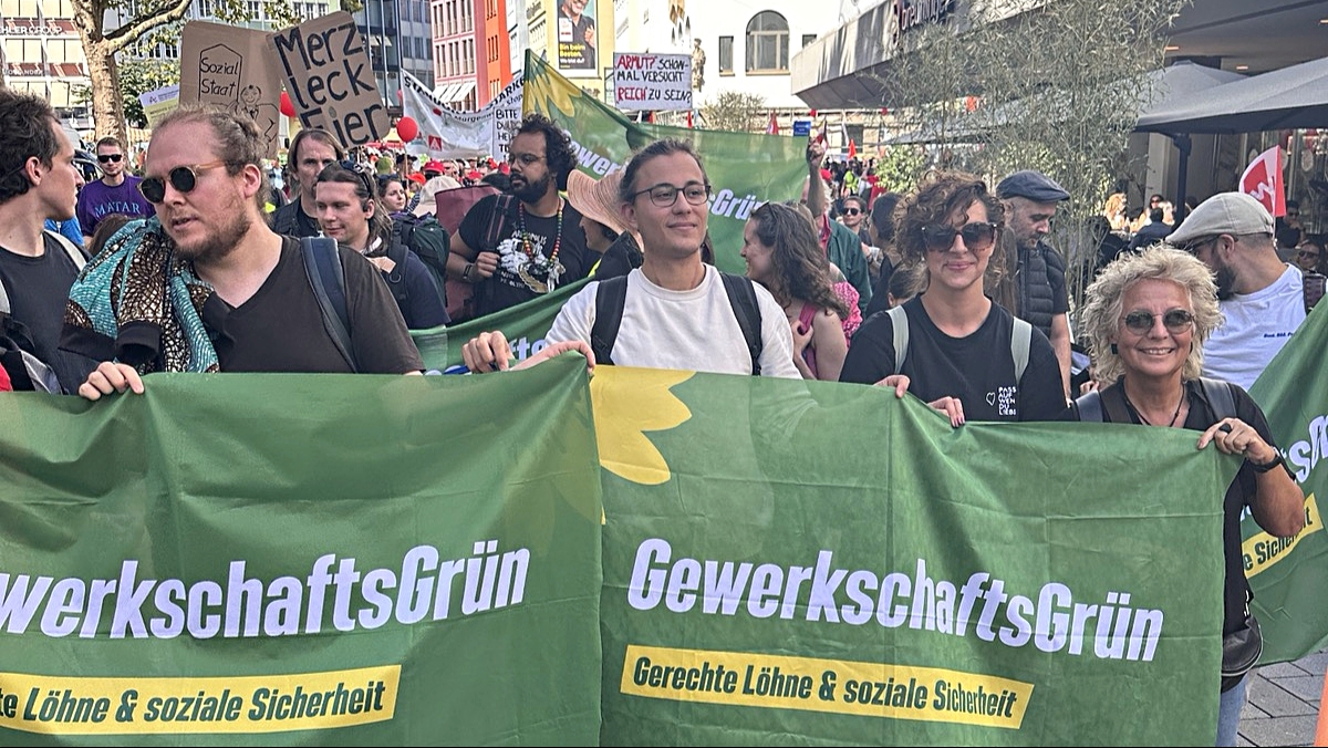Demonstrationszug mit GewerkschaftsGrün inmitten vieler Teilnehmender. Im Vordergrund werden zwei große grüne GewerkschaftsGrün-Banner getragen, im Hintergrund sind weitere Fahnen und Protestschilder zu sehen.