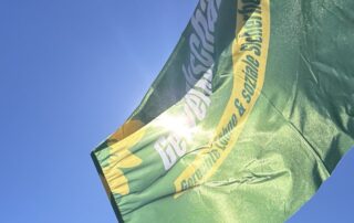 Grüne Fahne von GewerkschaftsGrün weht vor blauem Himmel. Auf der Fahne sind der Schriftzug GewerkschaftsGrün und „Gerechte Löhne & soziale Sicherheit“ zu erkennen.