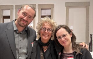 Beate Müller-Gemmeke steht beim BA-Empfang im Bundestag zwischen zwei ehemaligen Mitarbeiter:innen. Alle drei lächeln in die Kamera.