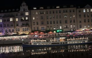 Nächtlicher Blick auf die Spree in Berlin. Ein beleuchtetes Ausflugsschiff fährt auf dem Wasser, dahinter liegen Restaurants und Häuser mit vielen Lichtern, die sich im Fluss spiegeln.