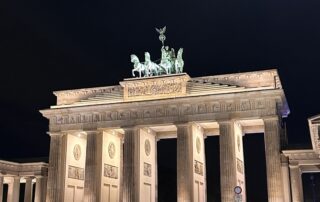 Das Brandenburger Tor in Berlin bei Nacht. Das Tor und die Quadriga sind hell angestrahlt, davor sind einige Menschen und ein Fahrradfahrer zu sehen.