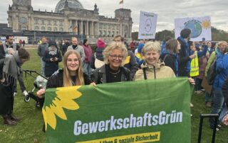 Beate Müller-Gemmeke steht mit Teilnehmenden der Jahrestagung von GewerkschaftsGrün bei der Demonstration für die Erneuerbaren auf der Reichstagswiese in Berlin. Sie halten gemeinsam ein großes GewerkschaftsGrün-Banner vor dem Reichstagsgebäude.