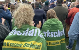 Beate Müller-Gemmeke steht mitten in der Demonstration für die Erneuerbaren in Berlin und trägt ein GewerkschaftsGrün-Banner über den Schultern. Vor ihr stehen zahlreiche weitere Demonstrierende mit Plakaten.