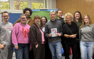 Gruppenfoto beim Austausch zwischen GewerkschaftsGrün und der Grünen Jugend auf der Jahrestagung in Berlin-Wannsee. Beate Müller-Gemmeke steht mit Vertreter beider Gruppen vor einem GewerkschaftsGrün-Banner. Ein Teilnehmer hält eine Broschüre der Initiative Aktion Wind in den Händen.