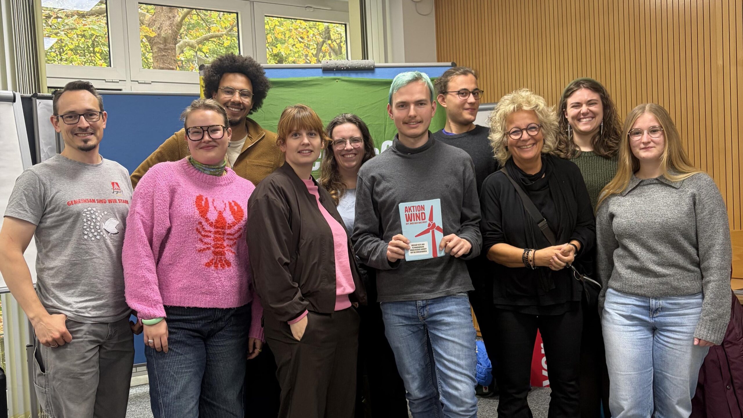 Gruppenfoto beim Austausch zwischen GewerkschaftsGrün und der Grünen Jugend auf der Jahrestagung in Berlin-Wannsee. Beate Müller-Gemmeke steht mit Vertreter beider Gruppen vor einem GewerkschaftsGrün-Banner. Ein Teilnehmer hält eine Broschüre der Initiative Aktion Wind in den Händen.